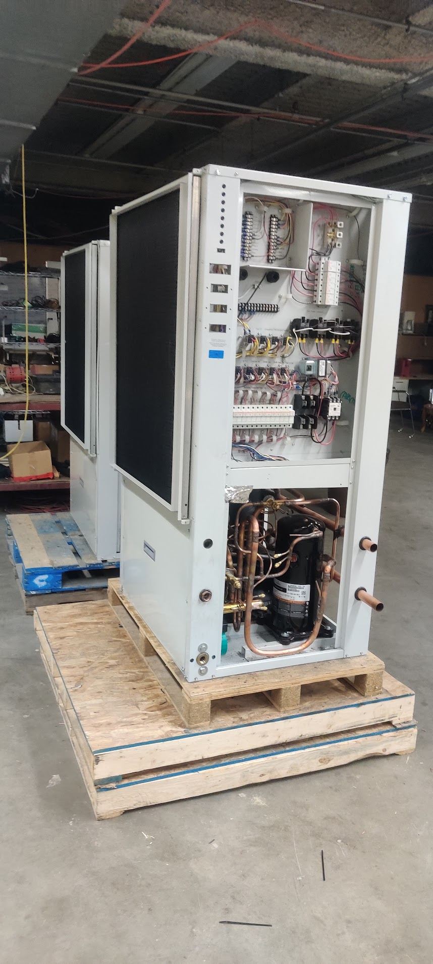 Two air-cooled condensing units on pallets in the shop, one with its electrical panel open showing breakers, contactors and wiring.