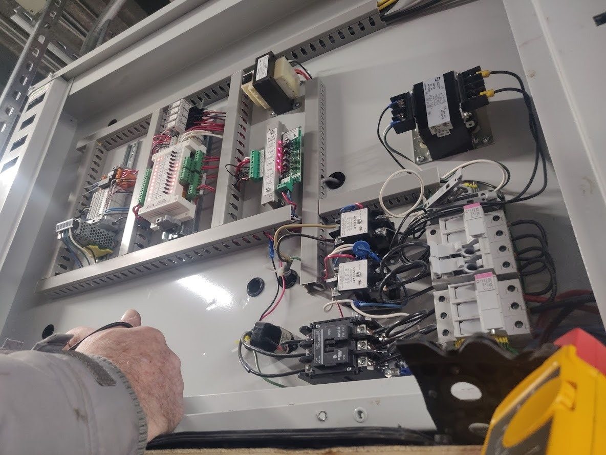 A technician working inside an open control panel: contactors, breakers, a transformer and labelled terminal strips.